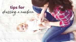 A mother kneels beside her infant who is laying on a blanket while she snaps the babys onsie closed